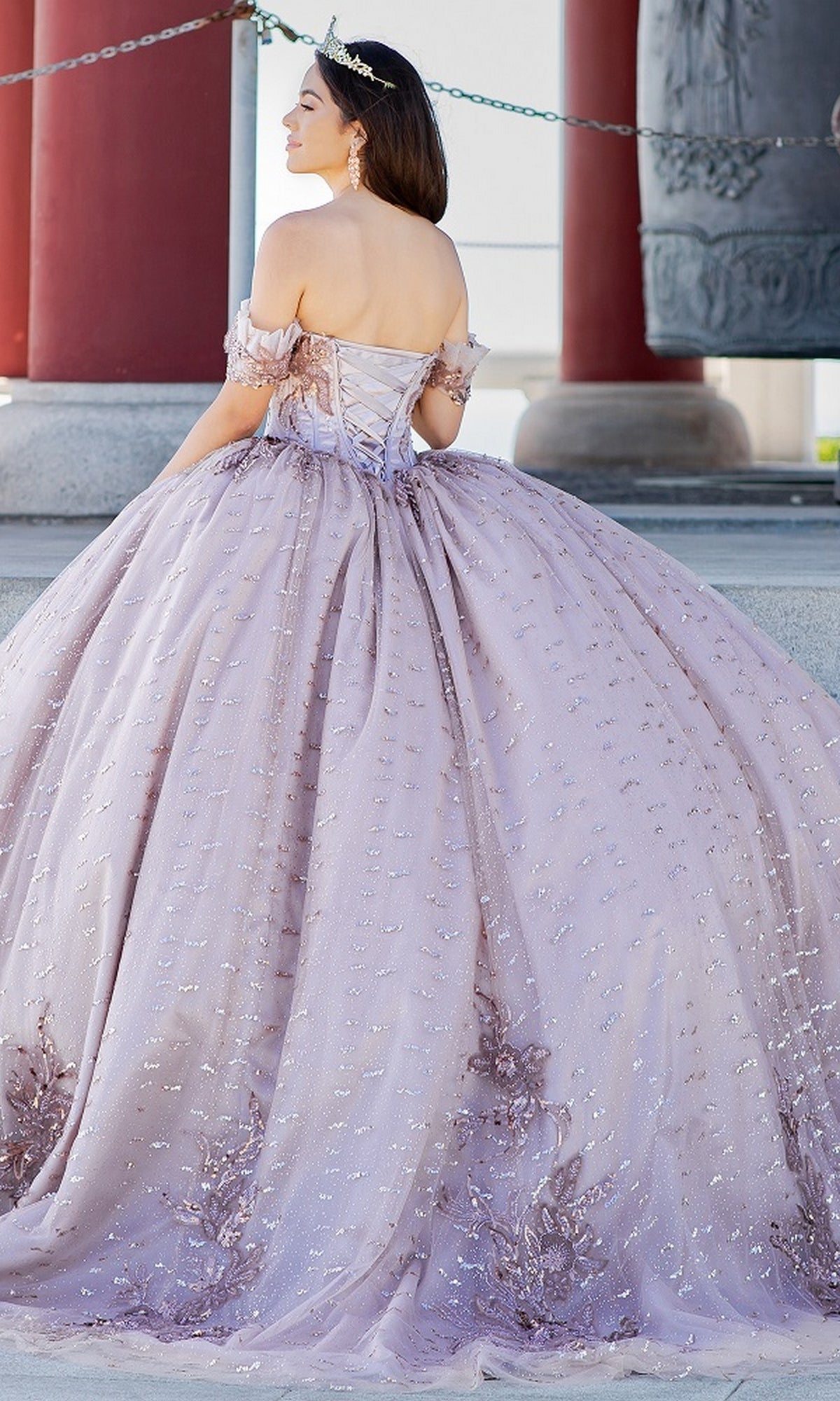 A woman in the Quinceanera Ball Gown 8087J, a light purple off-the-shoulder dress with a glitter tulle full skirt and tiara, stands outdoors facing away from the camera.
